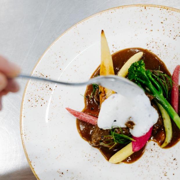 „Hände setzen den letzten Schliff an ein kunstvoll angerichtetes Gourmetgericht mit Sauce, Gemüse und Kräutern auf weißem Teller.